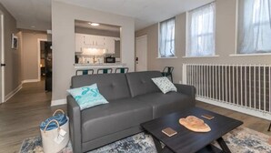 A modern living room with a grey sofa, a coffee table, and a kitchen area in the background. - Welcome to the Lakeside Suite! Enjoy! (Fairport Harbor)