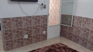 A bedroom with a stone-patterned wall, a flat-screen TV mounted above a door, and a fluffy brown rug on the floor. - Meu cantinho  (Guaratingueta)