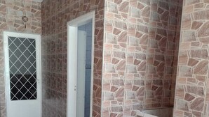 A room with stone-patterned walls, a tiled floor, a door with a metal grille, and a small shelf with a blue container. - Meu cantinho  (Guaratingueta)
