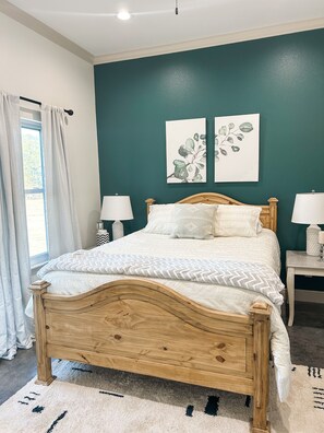 A bedroom with a wooden bed, two bedside tables with lamps, a window with curtains, and two wall-mounted artworks.