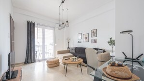 Ein modernes Wohnzimmer mit einer Couch, Couchtisch und Fernseher. Es gibt Korbnäpfchen, einen Glastisch und eine Stehlampe.