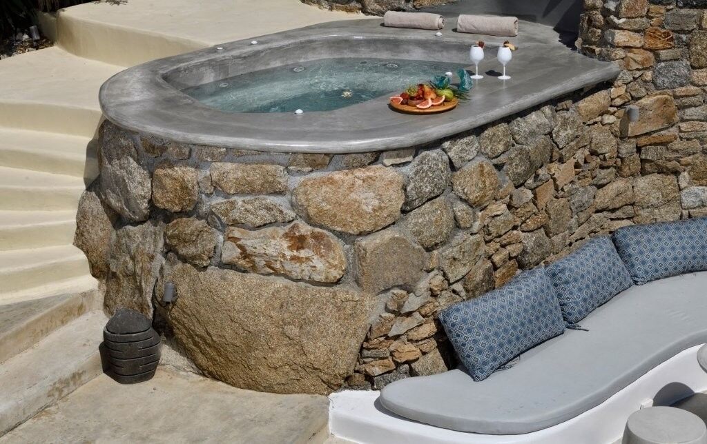 A stone hot tub with a wooden deck, a lounge chair, and a small table with a fruit platter.