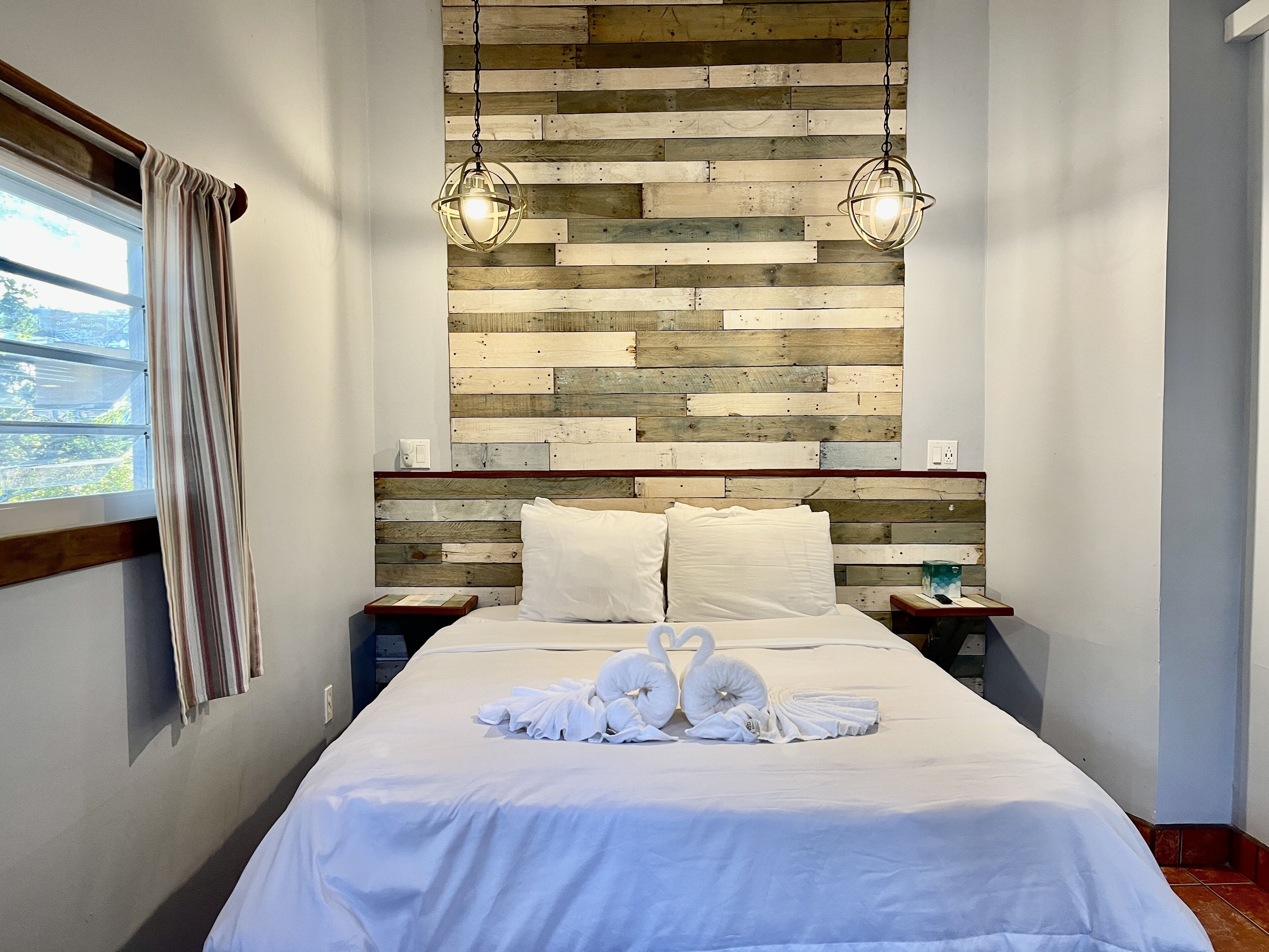 A neatly made bed with white linens and two swan-shaped towels, a wooden headboard with a rustic finish, and two pendant lights hanging above.