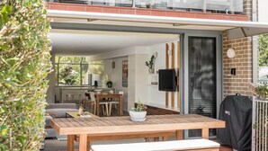 A modern kitchen with a dining area, a wooden table, and a grill outside. - 1 James Cook Apartments - Opposite Clarkes Beach (Byron Bay)