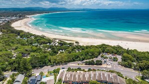 An aerial view of a coastal area with a beach, dense greenery, and a road with parked cars. - 1 James Cook Apartments - Opposite Clarkes Beach (Byron Bay)