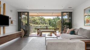 A modern living room with a flat-screen TV, a sofa, a coffee table, and a bench. There is a balcony with a view of greenery. - 1 James Cook Apartments - Opposite Clarkes Beach (Byron Bay)