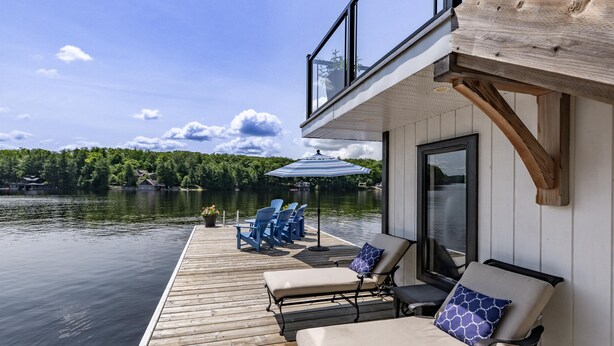 A wooden deck with lounge chairs and a blue umbrella, overlooking a lake with a forested shoreline. - All Day Sun! 7 bed 2 bath cottage on Lake Muskoka! Perfect Location! Privacy! (Torrance)