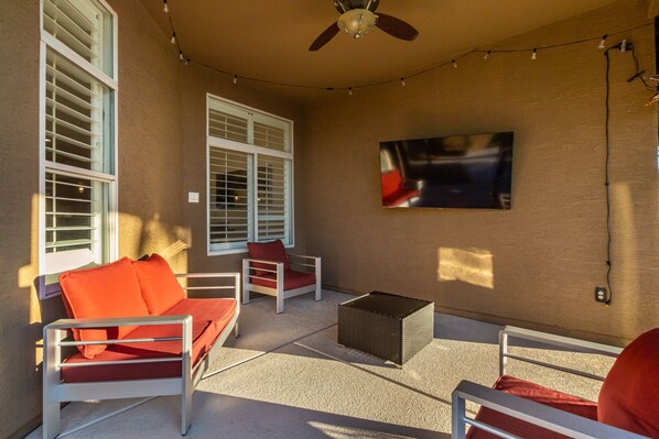 An outdoor patio with white furniture, a ceiling fan, string lights, and a TV mounted on the wall. - Phoenix Vacation Home -- Pool, Spa, Basketball & Putting Green (Phoenix)