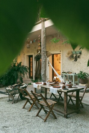 Un área de comedor al aire libre con mesas y sillas de madera rústica, rodeada de plantas en macetas y un muro de piedra.