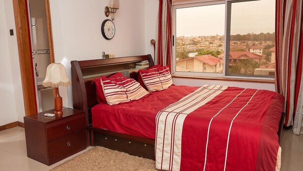 Een slaapkamer met een bed, nachtkastje, lamp, klok en een raam met uitzicht op gebouwen.