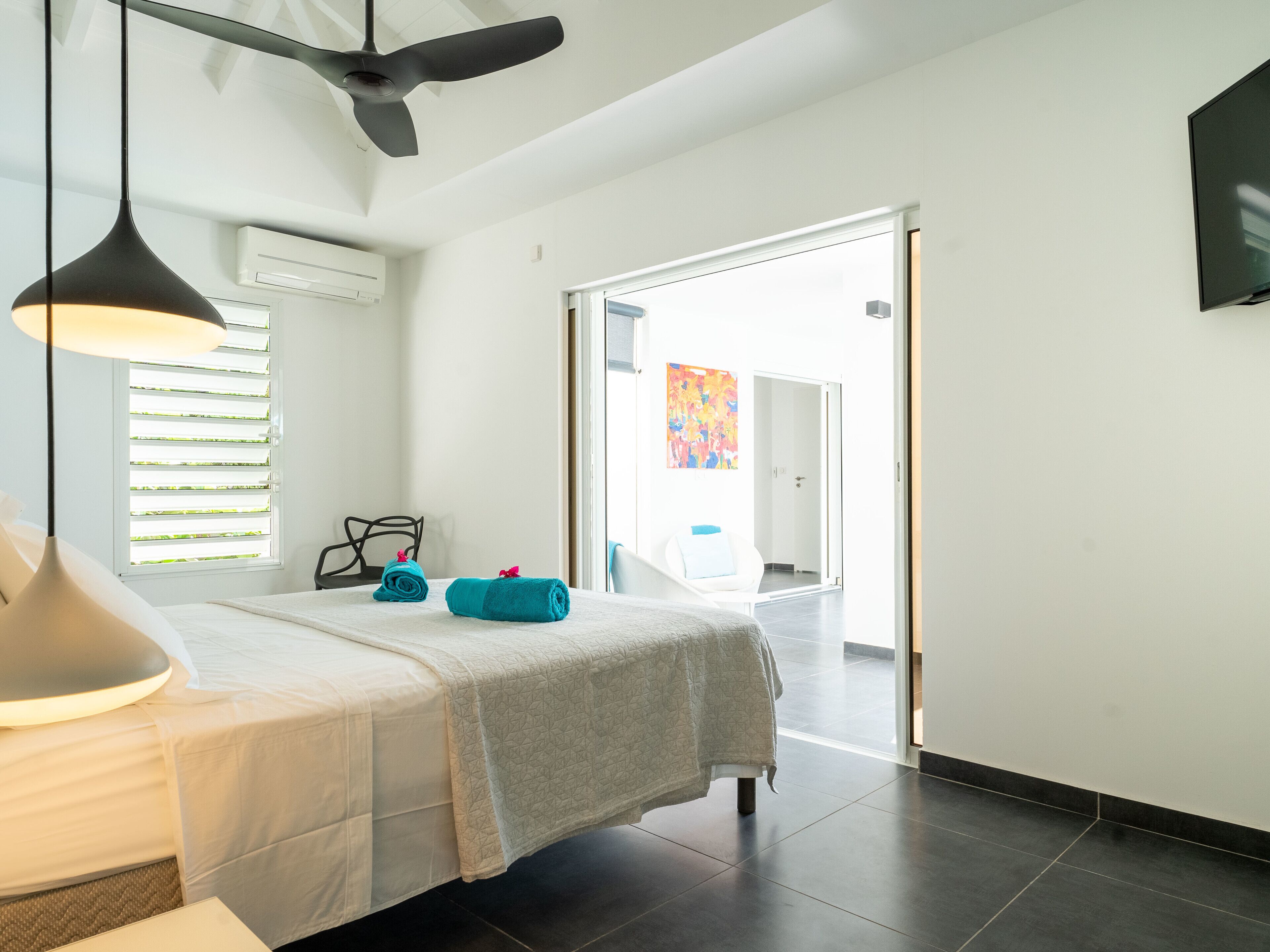 Una habitación de hotel moderna con una cama, un ventilador de techo, una ventana con persianas, un cuadro en la pared y vistas al pasillo.