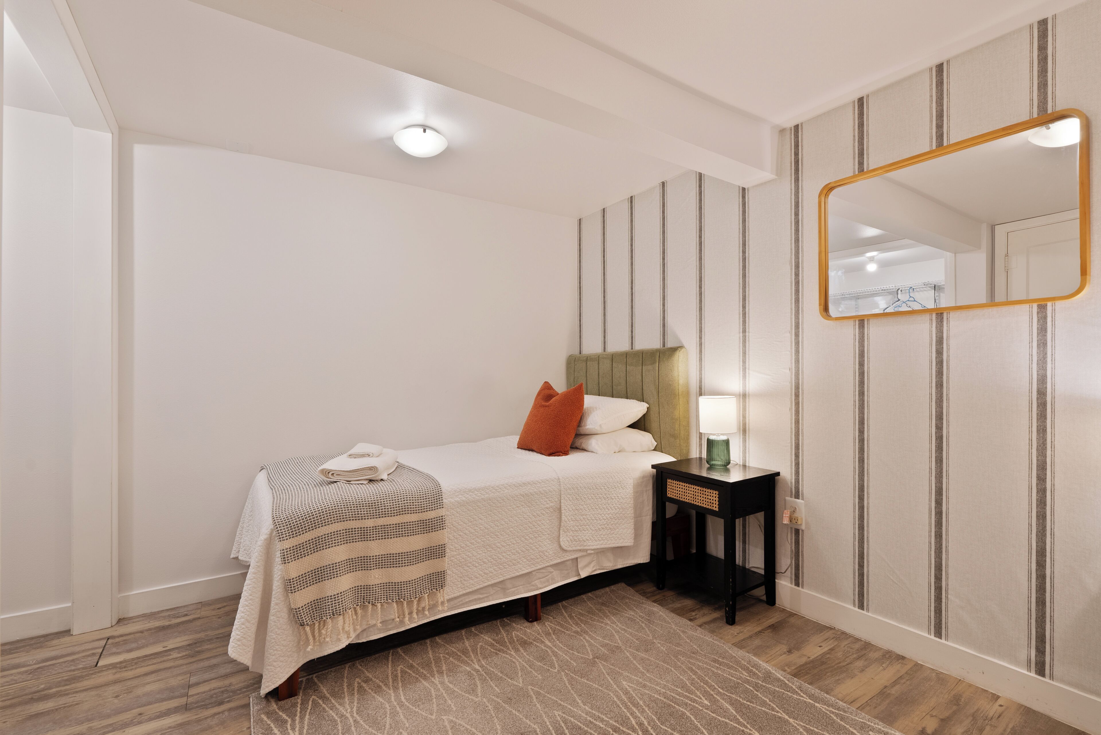 A neatly arranged bedroom with a bed, bedside table, lamp, and a mirror on the wall.