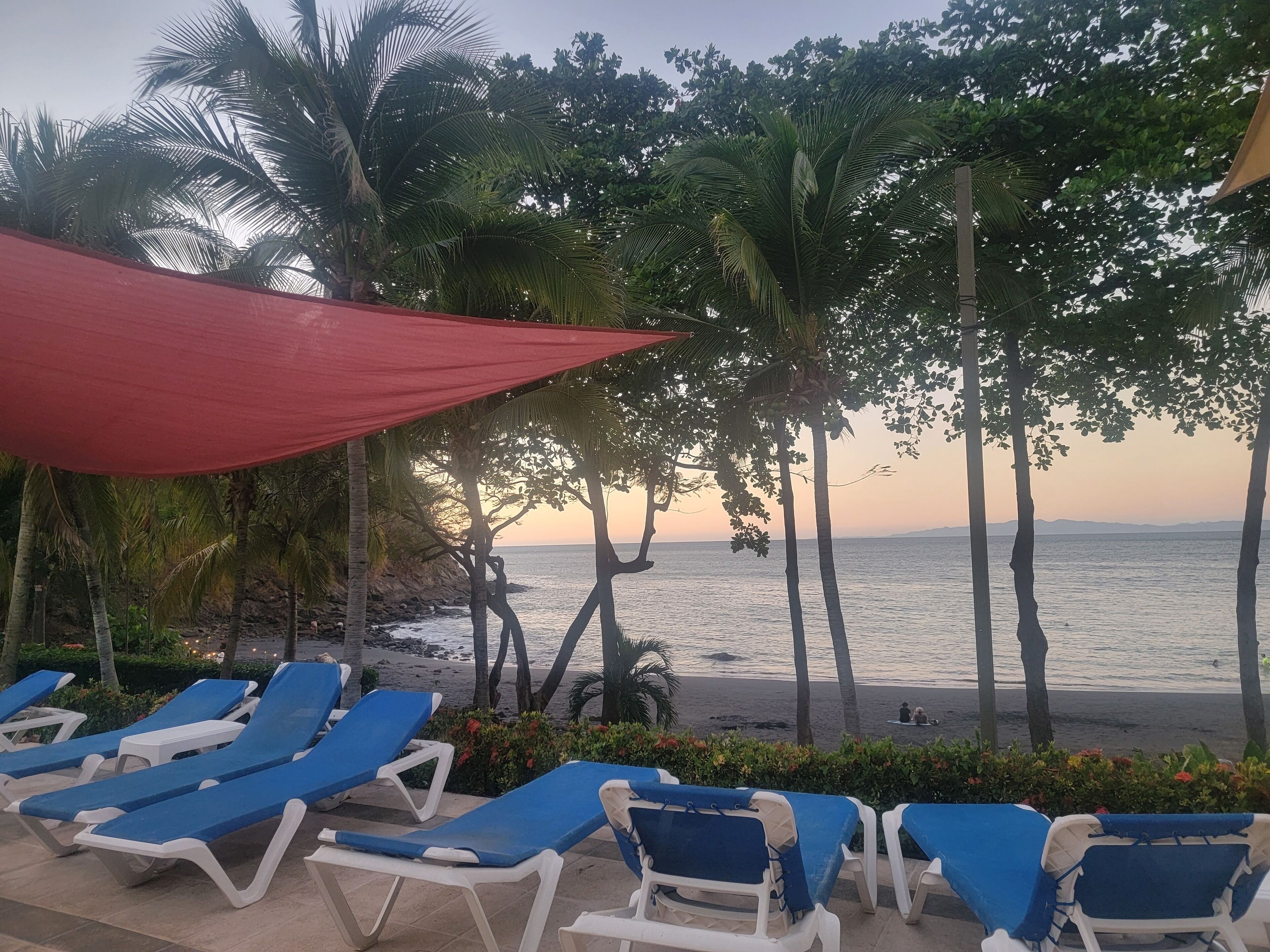 Un espace de détente aménagé sur la plage, avec des transats bleus et blancs, des palmiers et un auvent rouge.