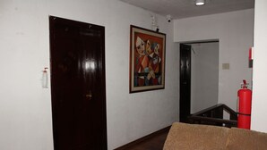 A hallway with a wooden door, a framed painting on the wall, a red fire extinguisher, and a brown sofa. - Hostal La Catolica (Quito)