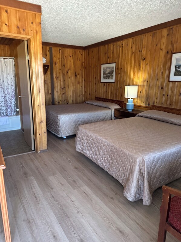 A room with wooden walls, two beds, a nightstand with a lamp, and a door leading to a balcony. - Glacier View Inn (Haines Junction)