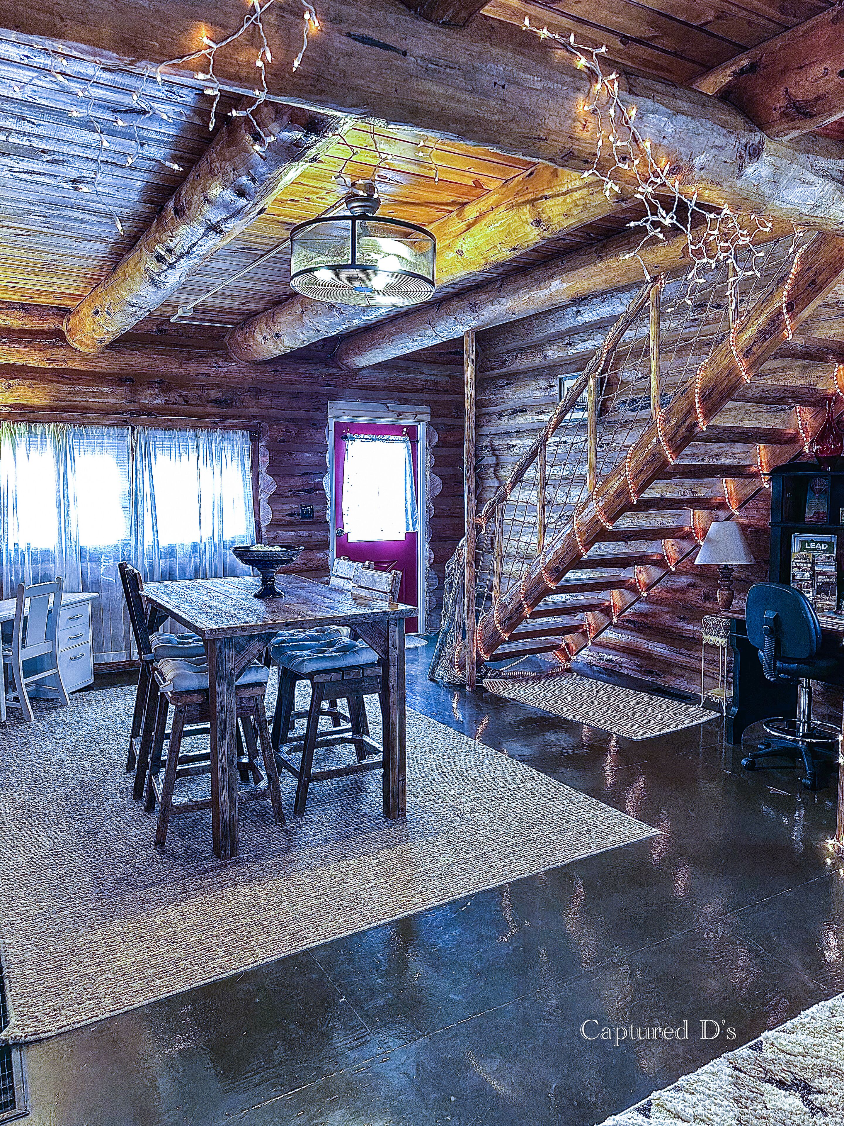 Interior rústico de cabana com mesa de jantar de madeira, cadeiras, escada e um pequeno espaço de escritório.