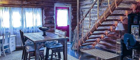 Interior rústico de cabana com mesa de jantar de madeira, cadeiras, escada e um pequeno espaço de escritório.
