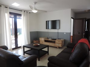 Una sala de estar moderna con un televisor de pantalla plana montado en la pared, un ventilador de techo, una mesa de centro con un frutero y un sofá.