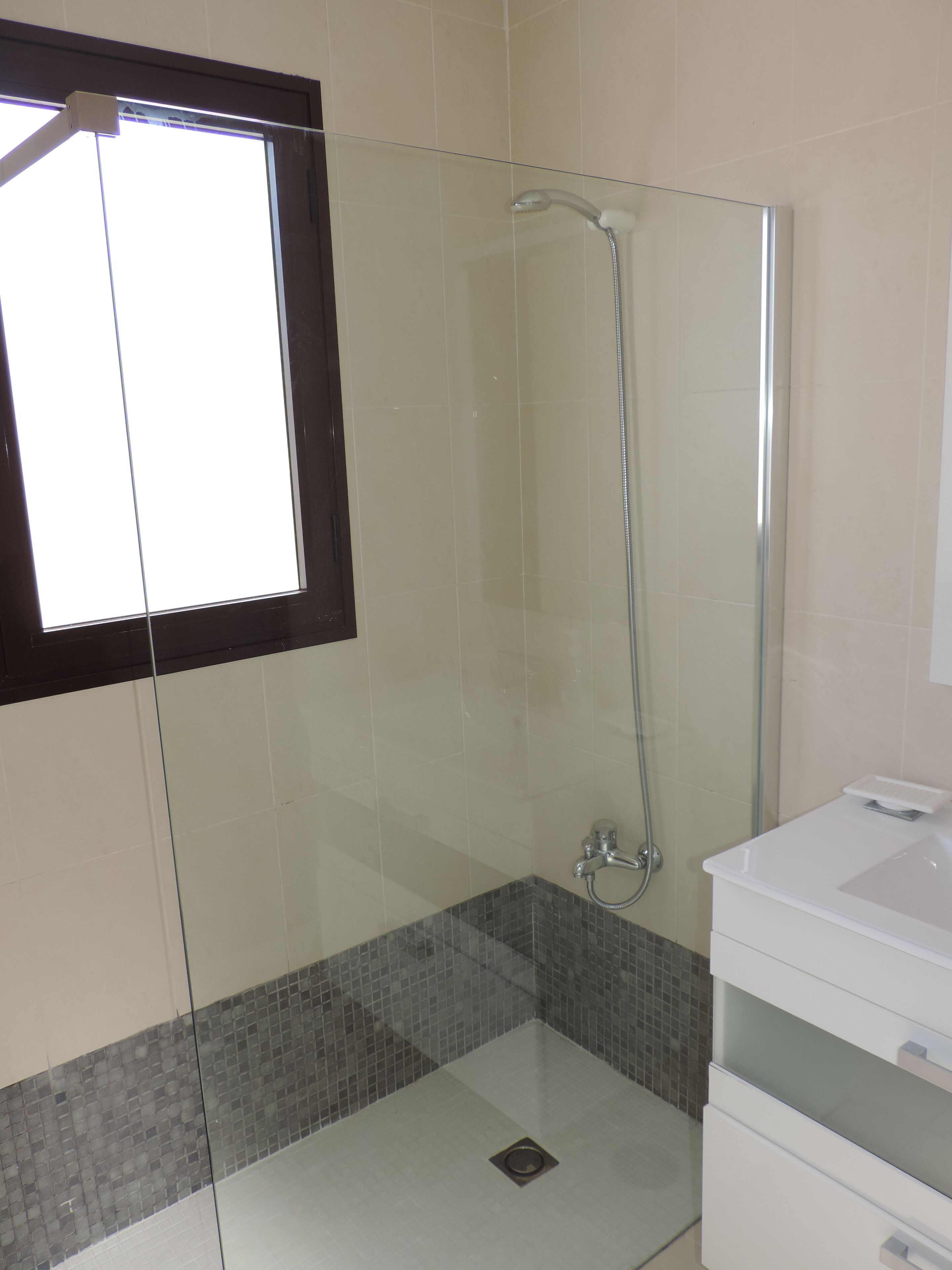 Zona de ducha con mampara de vidrio, cabezal de ducha manual, suelo de baldosas y lavamanos blanco con espejo.