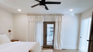 Une chambre spacieuse avec un grand lit, une chaise, un ventilateur de plafond et une vue sur l’extérieur, visible à travers une porte vitrée.