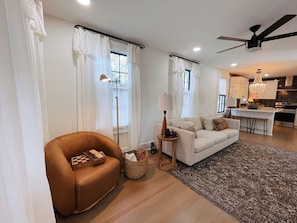 A modern living room with a sofa, armchair, and a chessboard. There is a lamp, a small table, and a rug. The room has large windows with curtains.