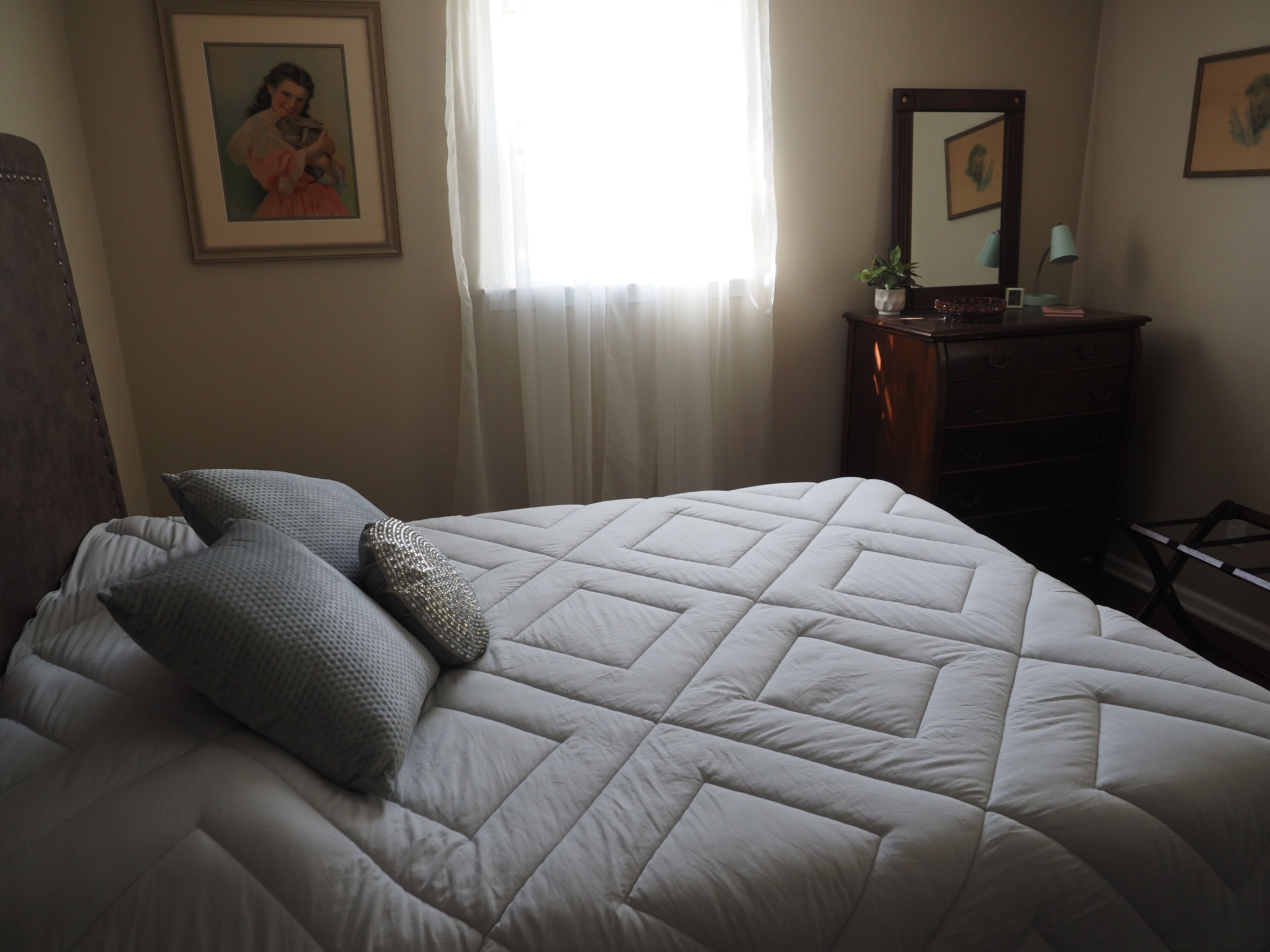 A neatly made bed with a quilted white duvet, two grey pillows, and a decorative pillow with a beaded pattern. There is a wooden dresser with a mirror above it, a small potted plant, and a framed picture on the wall.