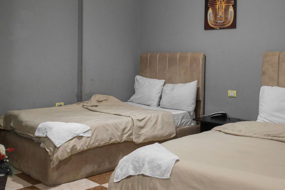 Une chambre d’hôtel avec deux lits, chacun recouvert d’une couvre-lit beige et agrémenté d’oreillers blancs. On y trouve une petite table avec un téléphone et une prise électrique murale.