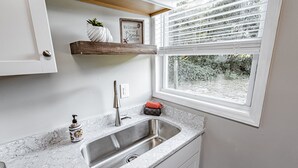 Una cocina con encimera de mármol, pileta de acero inoxidable, repisa de madera con adornos y una ventana con persianas.
