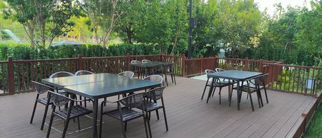An outdoor deck with two dining tables and chairs, surrounded by a wooden fence and lush greenery.
