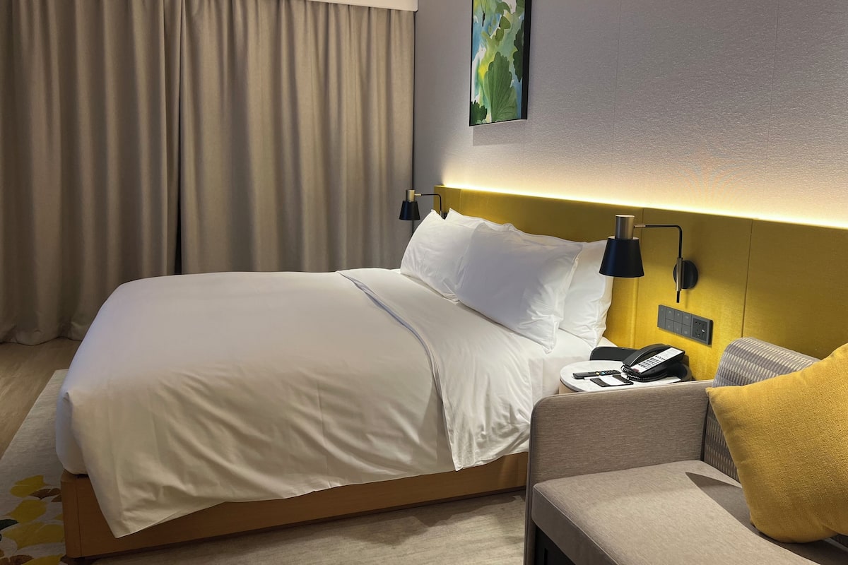 Quarto de hotel com uma cama, uma mesa de cabeceira com telefone, abajur e uma cadeira com almofada amarela.