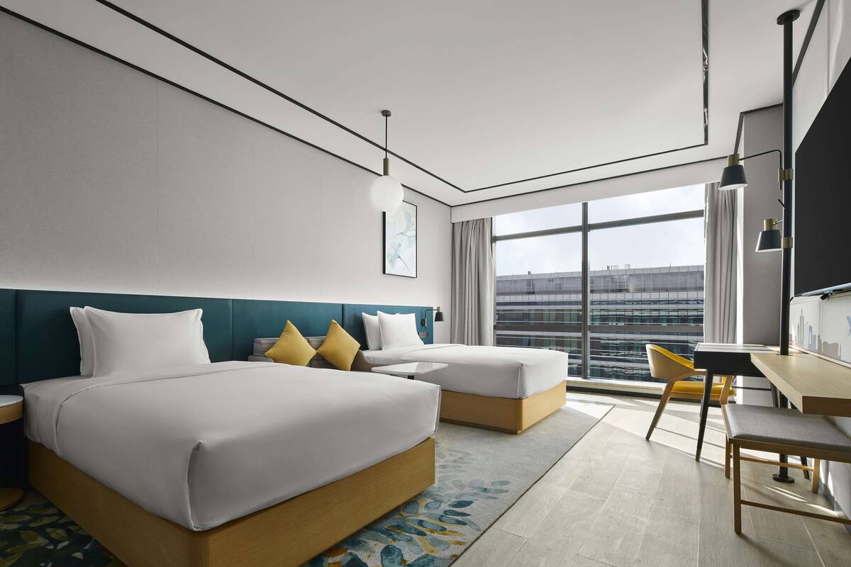 Quarto de hotel moderno com duas camas, uma escrivaninha e vista para a cidade.