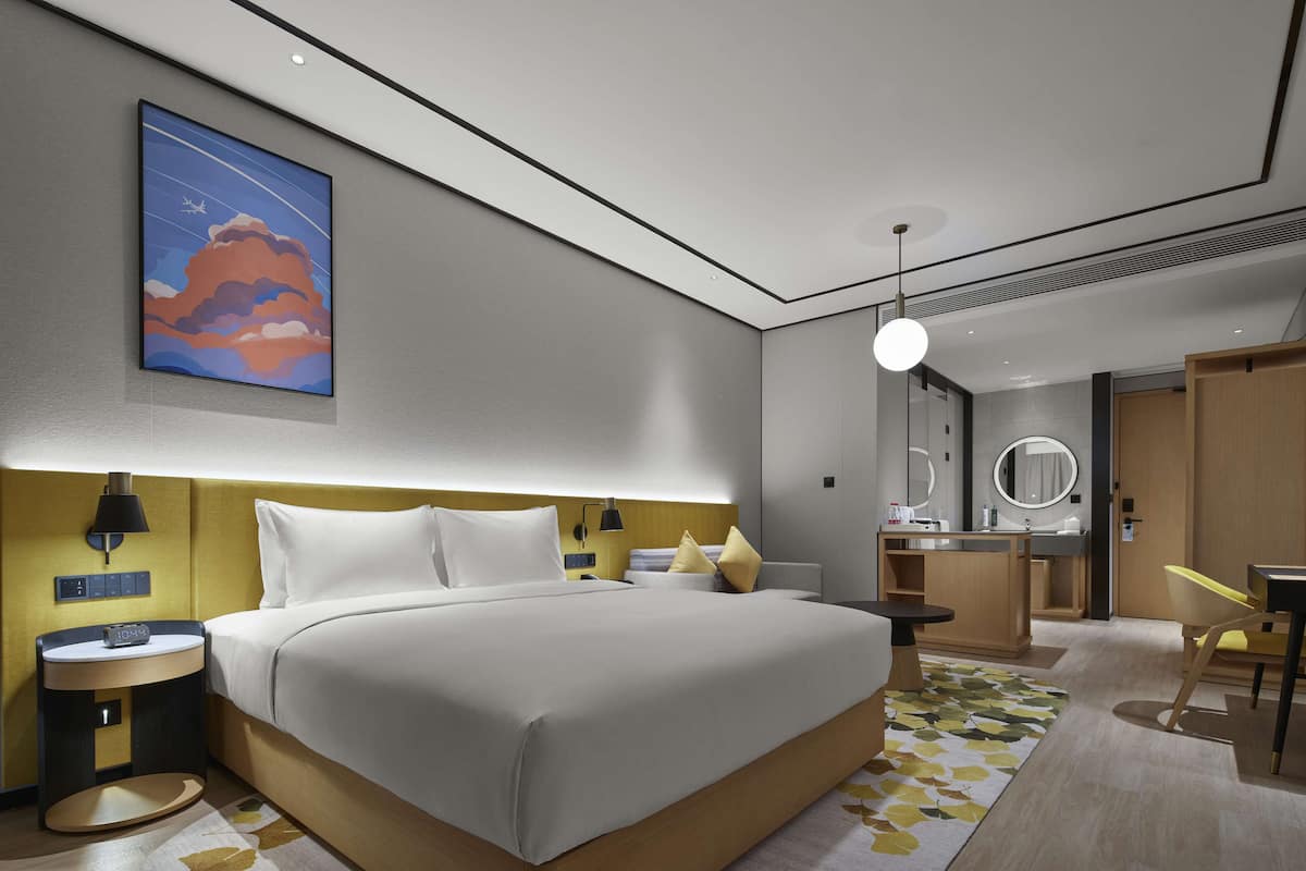 Quarto de hotel moderno com uma cama grande, mesa de cabeceira, abajur e banheiro com espelho e pia.
