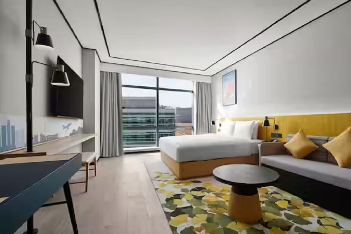A modern hotel room with a large bed, a sofa, a small table, a TV, and a balcony with a city view.