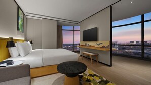 A modern hotel room with a large bed, a desk, a chair, a TV, and a city view.