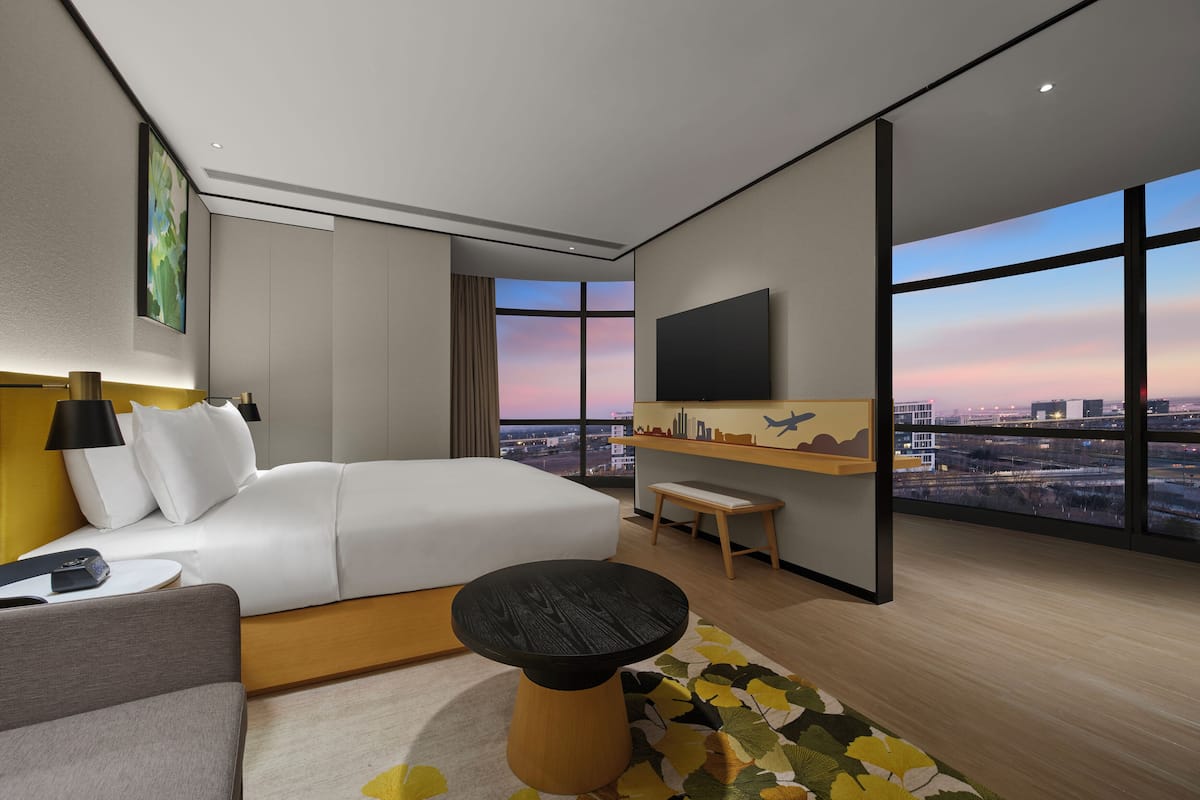 Quarto de hotel moderno com uma cama grande, uma escrivaninha, uma cadeira, televisão e vista para a cidade.