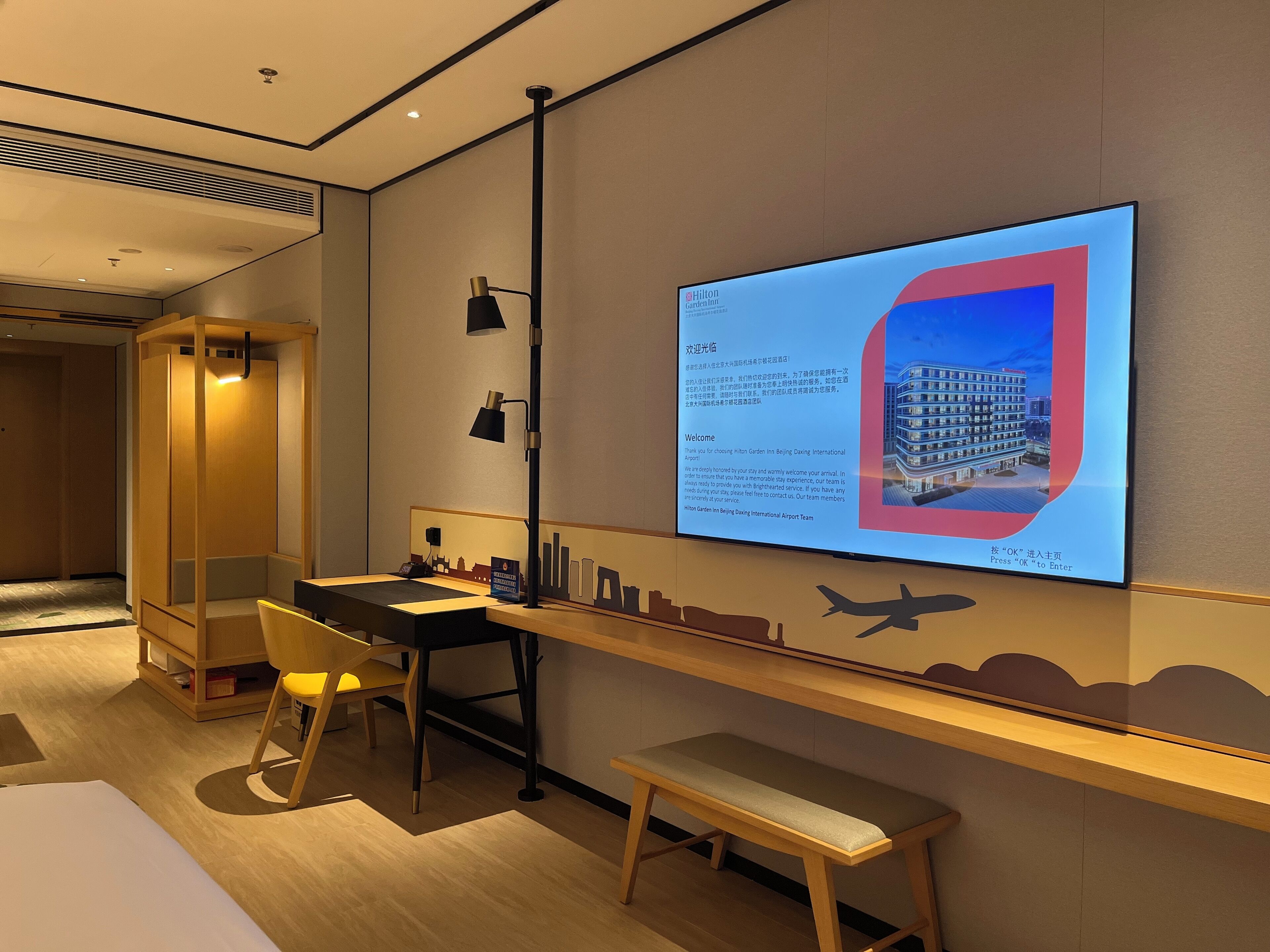 Ein Hotelzimmer mit Schreibtisch, Stuhl und Sitzbank. Ein großer Bildschirm zeigt eine Willkommensnachricht und ein Stadtbild.