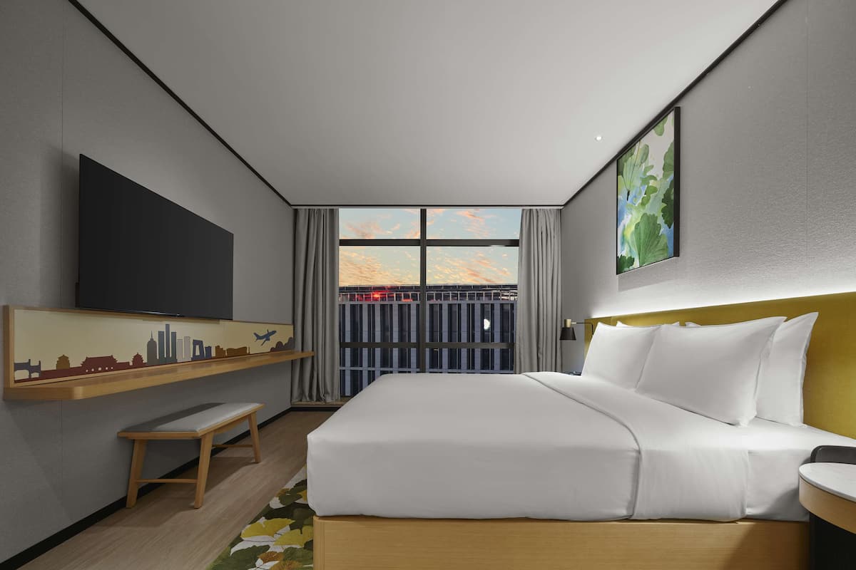 Quarto de hotel moderno com uma cama grande, televisão de tela plana, uma mesinha e uma obra de arte com vista para a cidade na parede.