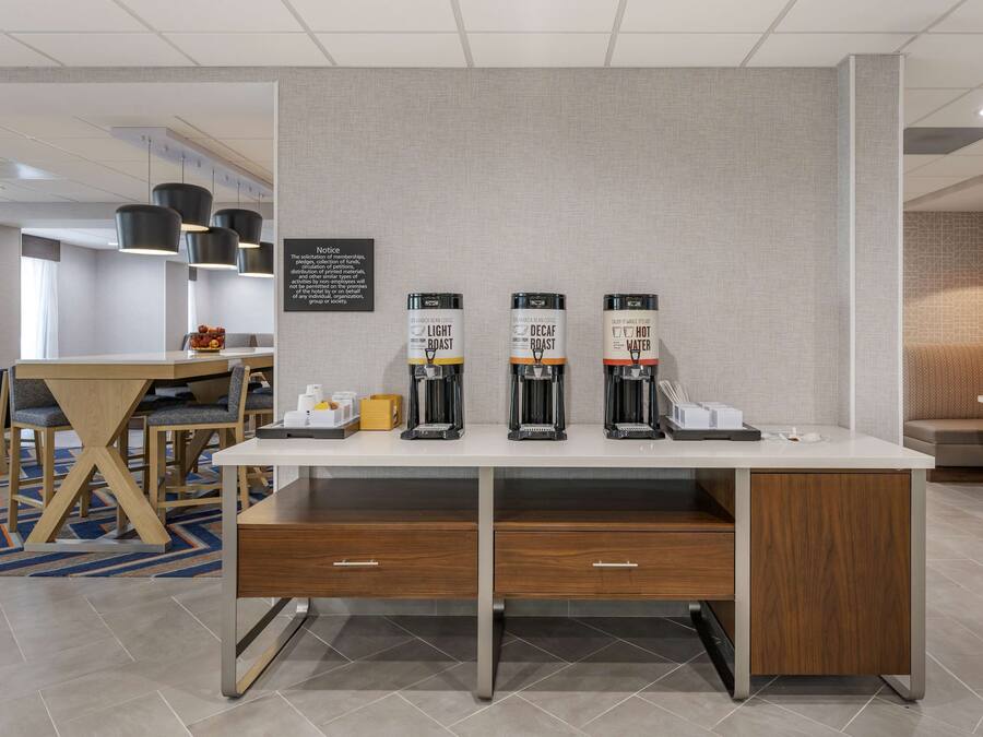 A hotel room with a white desk, three coffee machines, and a small table with chairs.