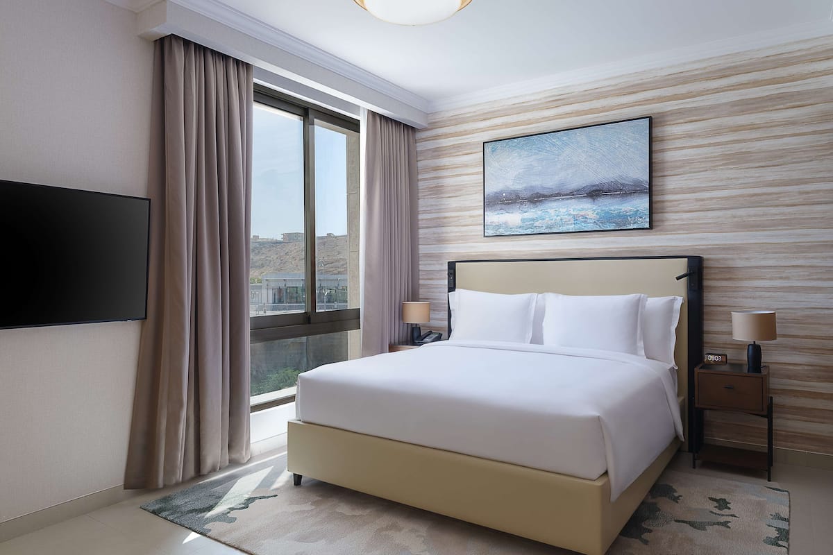 A hotel room with a large bed, a flat-screen TV mounted on the wall, a nightstand with a lamp, and a window with curtains.
