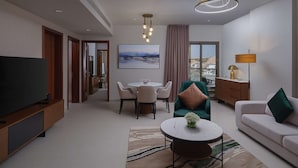 A modern hotel room with a flat-screen TV, a dining area with a table and chairs, a sofa with cushions, a coffee table with a vase, and a large window with a city view.