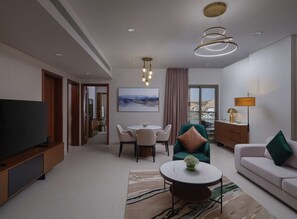 A modern hotel room with a flat-screen TV, a dining area with a table and chairs, a sofa with cushions, a coffee table with a vase, and a large window with a city view.