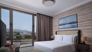 A hotel room with a large bed, a desk with a chair, a nightstand, and a painting on the wall.