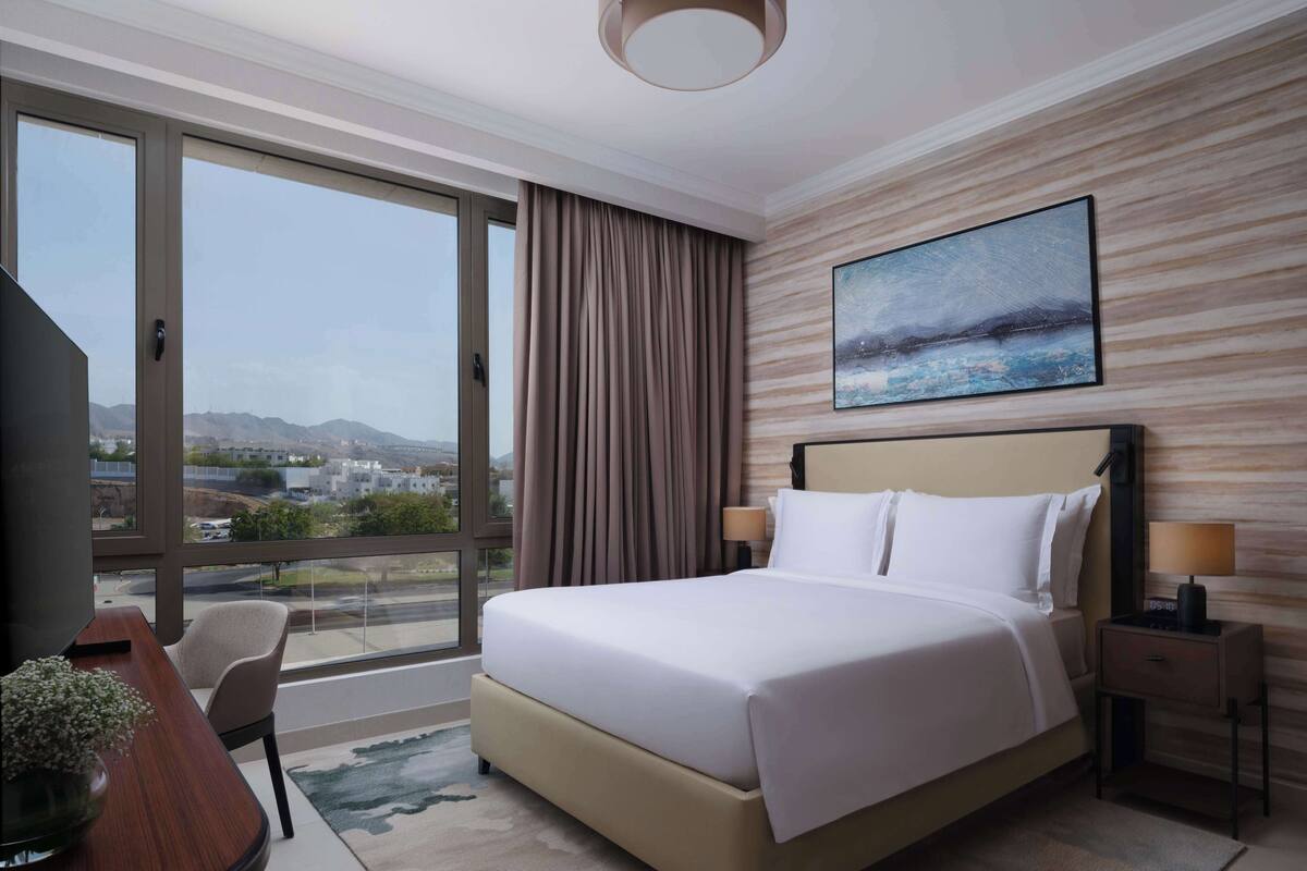 A hotel room with a large bed, a desk with a chair, a nightstand, and a painting on the wall.