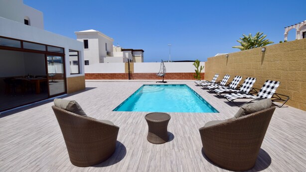 Zona de piscina con sillas de descanso y una mesa, rodeada de edificios modernos.