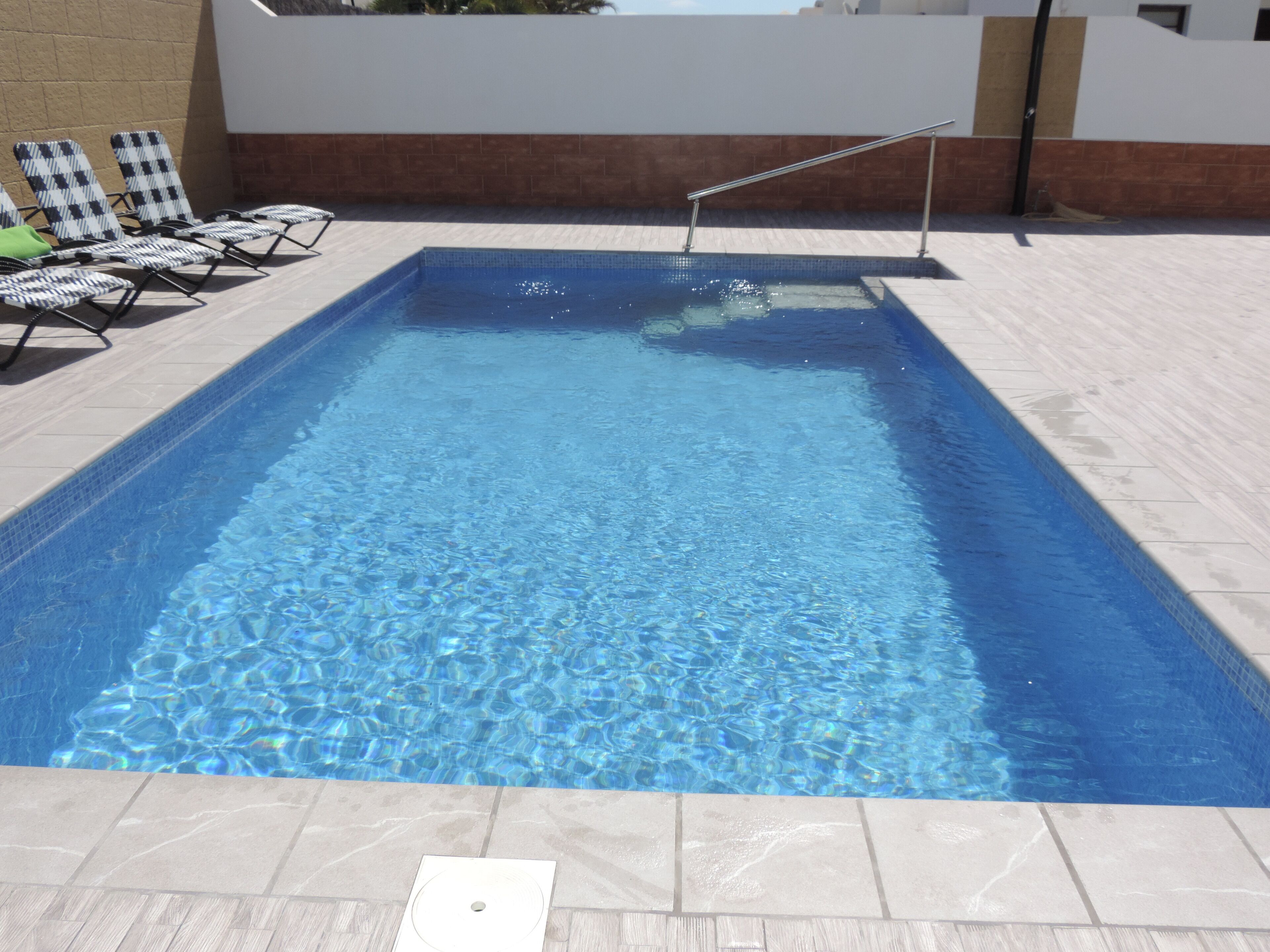 Ein rechteckiger Swimmingpool mit klarem, blauem Wasser, umgeben von hellgrauen Fliesen und einer niedrigen Mauer.