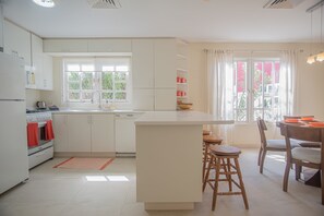 Uma cozinha com armários brancos, uma ilha central com banquetas, uma mesa de jantar com cadeiras e uma janela com cortinas.