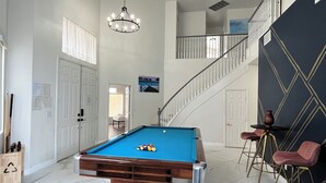 A pool table in a spacious room with a high ceiling, a staircase, and a view of the outdoors.