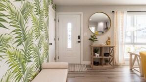 A room with a bed, a wooden floor, a white door, a mirror, a shelf with plants, and a window with curtains.
