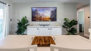 A dining area with a wooden table, white chairs, a white cabinet, and a large landscape painting on the wall.