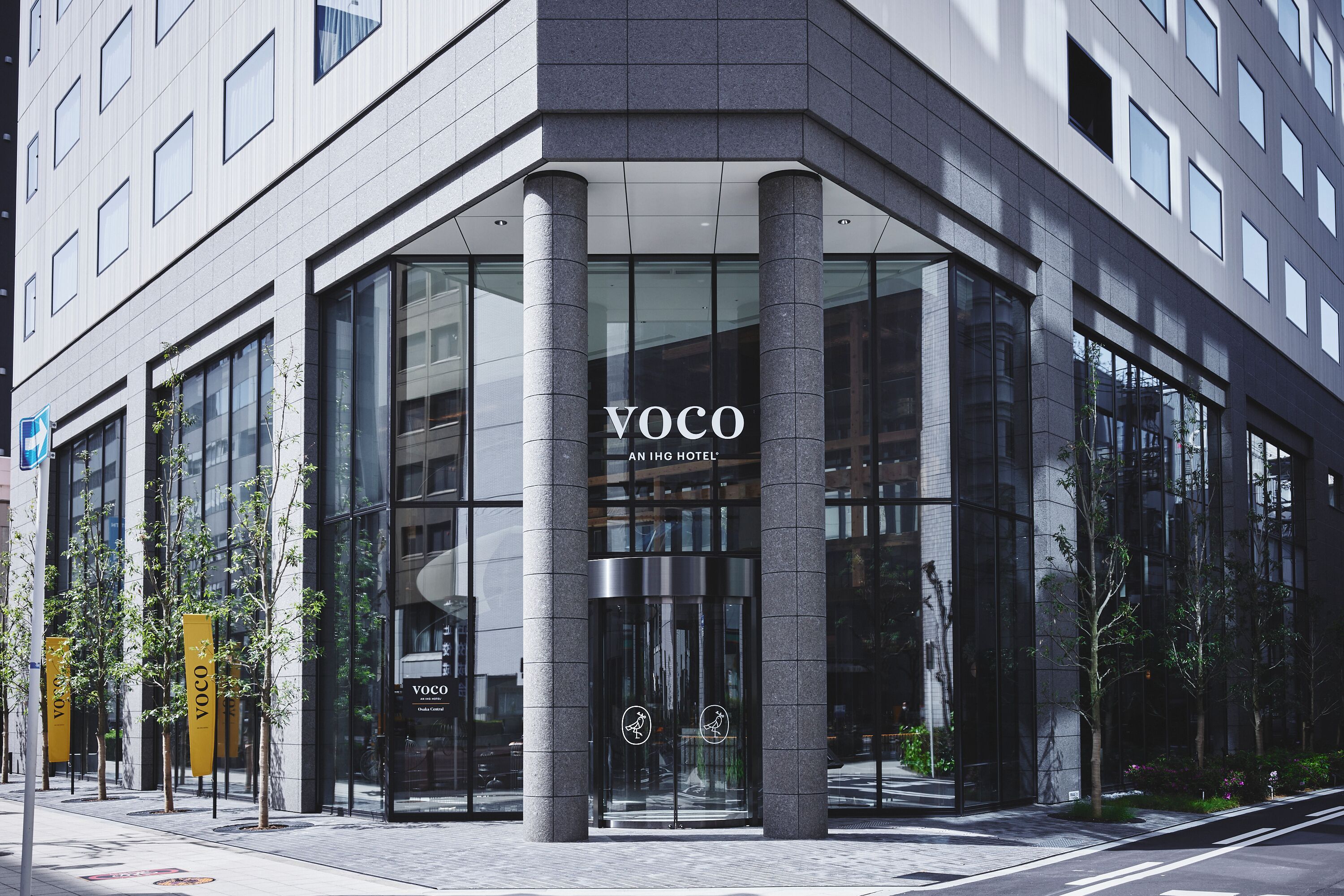 Une entrée d’hôtel moderne, avec des portes en verre, un logo bien visible et une vue dégagée sur la rue.